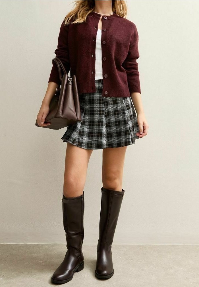 Femme portant un cardigan bordeaux, un haut blanc, une jupe à carreaux grise, des bottes marron montant jusqu'aux genoux et tenant un sac à main marron assorti.