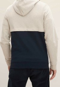 Uomo che indossa un hoodie bicolore con la parte superiore beige e la parte inferiore blu navy, abbinato a pantaloni scuri, visto da dietro.