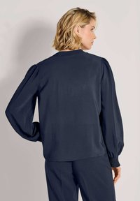 Frau mit kurzem blonden Haar, die eine marineblaue Bluse mit langen Ärmeln, Puffärmeln und gerafften Bündchen trägt und von der Kamera abgewandt ist.