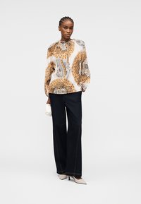 Femme debout portant un chemisier à manches longues beige et noir à motifs, un pantalon large noir, des talons blancs, et tenant une petite pochette blanche.