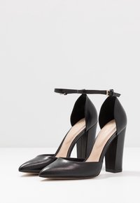 Černé kožené lodičky s špičatou špičkou, páskem kolem kotníku, otevřenými stranami a masivním čtvercovým podpatkem. Vnitřek má tělovou stélku.