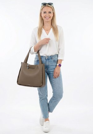 Blonde woman wearing white blouse, light blue jeans, white sneakers, holding large brown leather handbag with zipper and keychain detail.