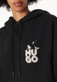 Svart hettegenser med snøring, prydet med et slitt grafisk motiv av "HUGO" omringet av sommerfugler i nyanser av beige og hvitt.