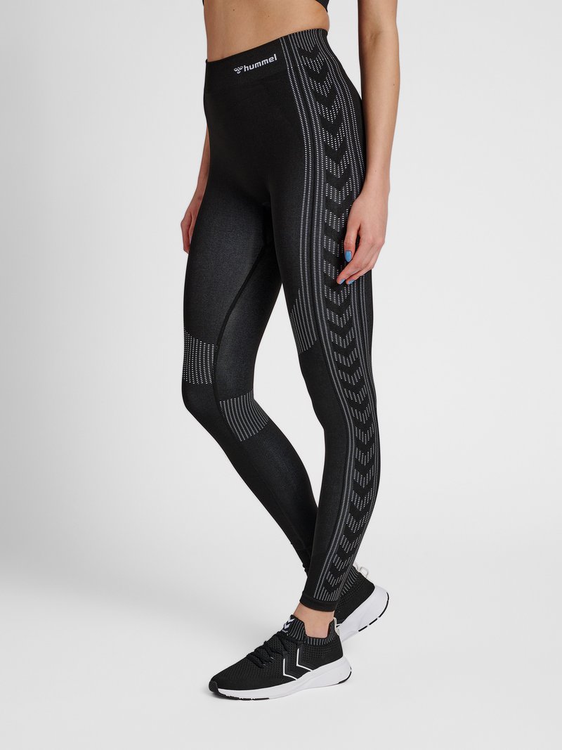 Kvinde iført sorte højttaljede leggings med mønstrede striber og sorte træningssko, stående mod en ensartet lys baggrund.