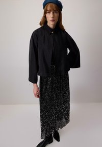 Woman wearing a black beret, black cropped jacket with gold button, black patterned pleated skirt, and black ankle boots, standing against neutral background.