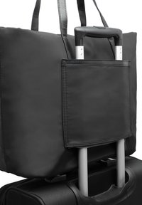 Bolso de viaje negro tipo tote sujeto al asa de una maleta negra vertical con ruedas, con el asa telescópica extendida.