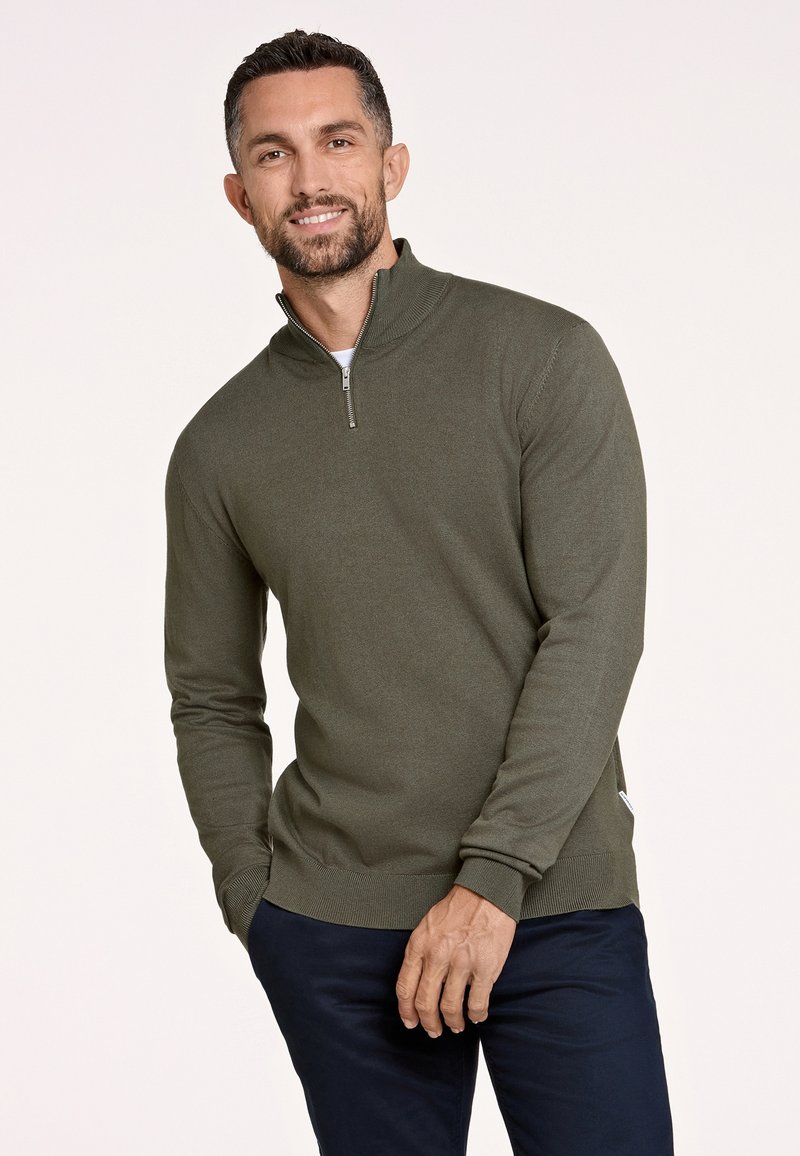 Olijfgroen sweater met ritssluiting en lange mouwen, gemaakt van zacht breiwerk en voorzien van geribbelde boorden, gecombineerd met marineblauwe pantalon.