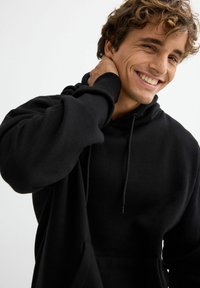 Sudadera con capucha negra hecha de tela suave con un bolsillo canguro, cordón ajustable y puños acanalados. El modelo sonríe, mostrando el diseño.