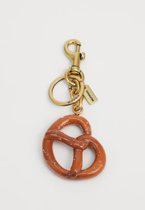 Bruine pretzelvormige sleutelhanger met een glanzende afwerking en witte zoutkorrels. Gouden metalen musketonhaak en ringen, met een merklabel eraan bevestigd.