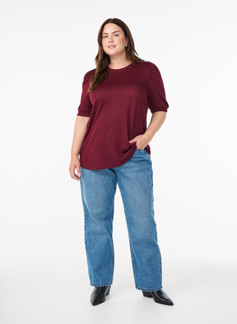 Vrouw draagt een bordeauxrode top met korte mouwen, blauwe spijkerbroek en zwarte schoenen, staat met één hand in de zak op een witte achtergrond.
