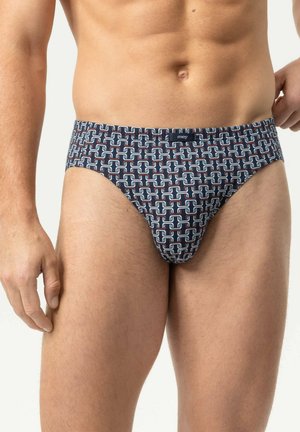 Männlicher Torso, der dunkelblaue Slips mit weißem und rotem geometrischem Muster trägt, vor einem einfarbigen hellen Hintergrund stehend.