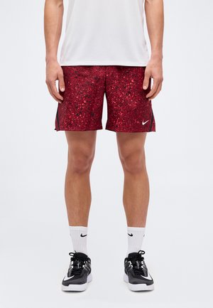 Mann trägt rote Nike-Shorts mit Sprenkeln, weiße Nike-Socken, schwarz-weiße Nike-Sneaker und ein weißes Hemd und steht vor schlichten Hintergrund.