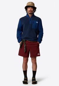 Mann iført beige bøttehatt, blå fleecepullover, burgunder shorts med grønt bandana, svarte sokker og sandaler, står mot hvit bakgrunn.