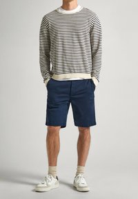 Un homme porte un pull à rayures beige et noir à manches longues, un short bleu marine, des chaussettes beiges et des baskets blanches, se tenant les mains dans les poches.