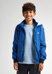 Jachetă albastră cu fermoar și glugă, având un model geometric subtil, manșete elastice și o patch logo mic. Purtați peste un tricou bleu deschis.