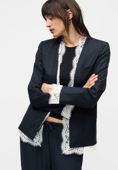 Γυναίκα με σκούρα μαλλιά μέχρι τους ώμους, που φορά μαύρο σακάκι με λευκή δαντέλα πάνω σε μαύρο τοπ και ταιριαστό μαύρο παντελόνι, με σταυρωμένα τα χέρια.