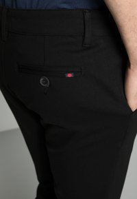 Black trousers featuring a back pocket with a button and a small red and navy tag accent. The fabric appears smooth with a tailored fit.