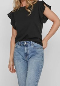 Černý top s krátkým rukávem a volánkovými akcenty, který je spárován se světle modrými vysokými džíny s knoflíkovým zapínáním a předními kapsami.