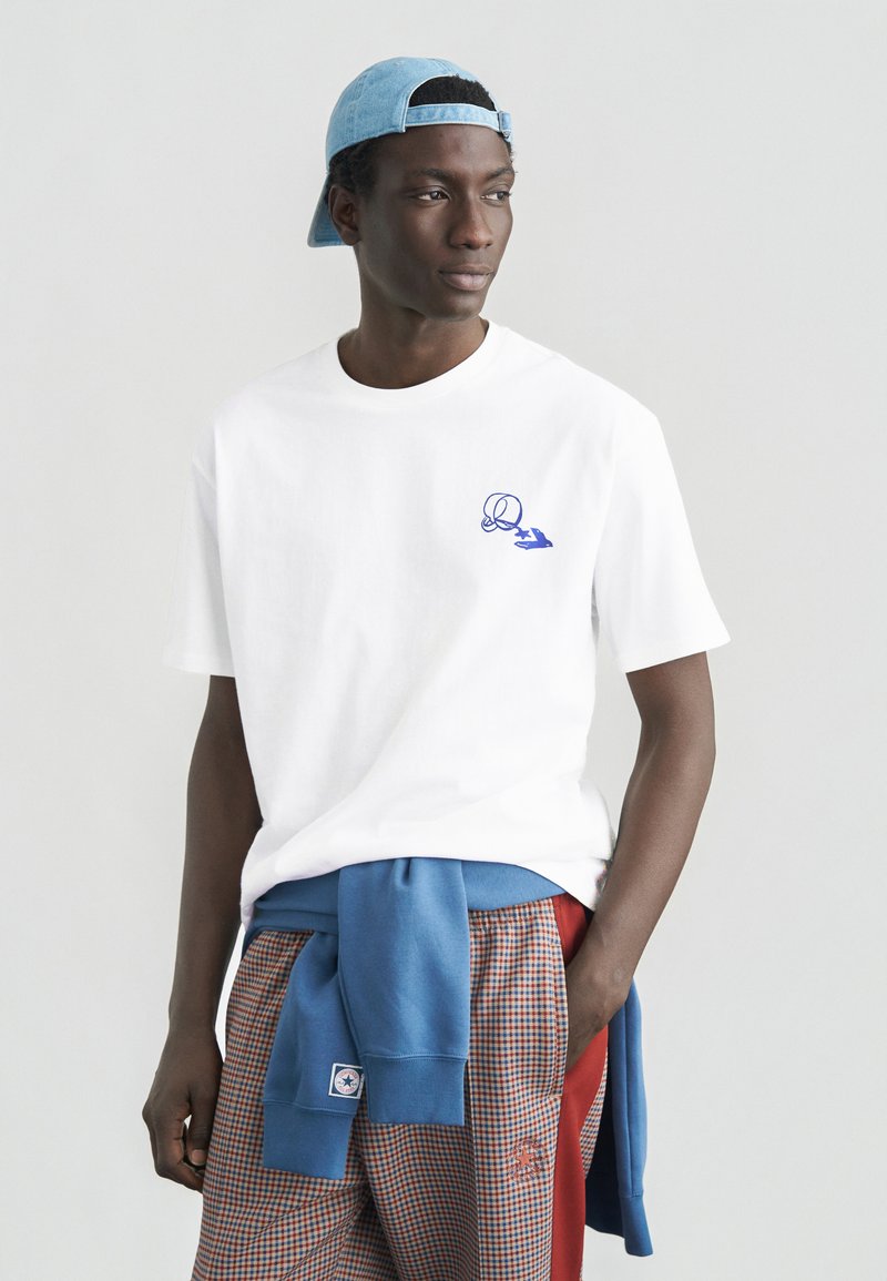 Λευκό βαμβακερό μπλουζάκι με μπλε σχέδιο στο στήθος, συνδυασμένο με παντελόνι με καρό και μπλε πουλόβερ δεμένο στη μέση.