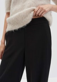 Une personne soulève un pull en laine beige et duveteux pour révéler un pantalon noir taille haute avec une ceinture lisse, le tout sur un fond uni.