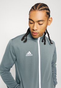 Grå zip-up jakke med hvide detaljer, lange ærmer og et Adidas-logo på venstre bryst. Lavet af glat, fleksibelt stof.