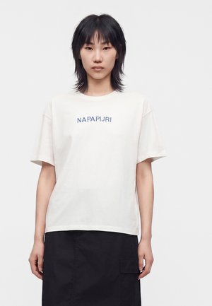 Ung person med kort sort hår iført en hvid Napapijri T-shirt og et sort skørt, stående mod en ensfarvet hvid baggrund.