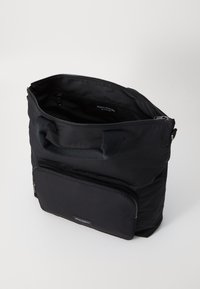 Sac fourre-tout en nylon noir avec deux poignées, poche zippée externe et fermeture éclair. Texture lisse, forme cubique, branding minimal à l'intérieur.