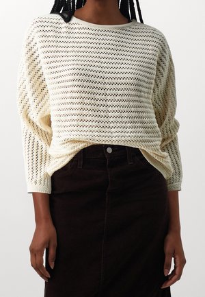 Personne portant un pull crème en maille ouverte à manches longues et une jupe noire taille haute avec une fermeture à boutons sur le devant.