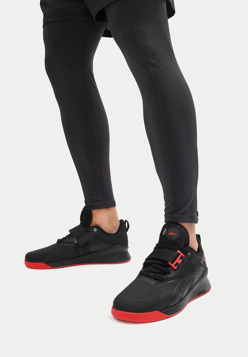 Czarne buty sportowe z czerwonymi akcentami, o smukłym designie, teksturowanymi overlayami i pojedynczym paskiem na środku stopy dla wsparcia.