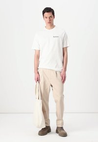 Witte katoenen t-shirt met "Barbour" logo, gecombineerd met beige geplooide broeken, bruine schoenen en een beige canvas draagtas.