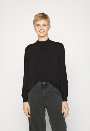 Black long-sleeve top with a high neckline and gathered detail at the collar; paired with high-waisted dark grey jeans. Smooth fabric texture.
