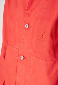 Koralinta jaka, kas izgatavota no viegla auduma, ar divām priekšējām kabatām, sudraba pogām un izšuvumu kreisajā krūtī.