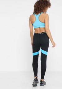 Sinivihreä urheilullinen lyhyt toppi ja mustat legginsit, joissa on siniset sivujuovat. Asusteet istuvat tiiviisti, ja niissä on saumaton muotoilu sekä suoritustason brändilogo.