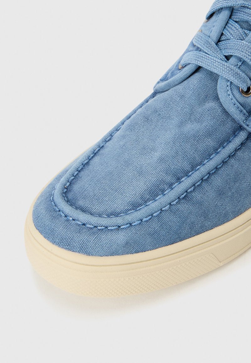 Zapatilla de lona azul con suela de goma beige, puntera redonda, costuras tonales y superficie texturizada. Presenta un cierre de cordones y un cuello acolchado.