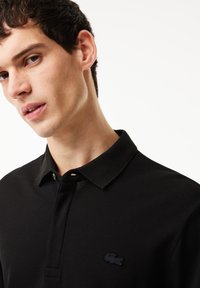Sort polo-shirt lavet af tekstureret stof, med en klassisk krave, to knapper og et lille broderet logo på brystet.