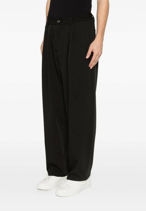 Uomo che indossa pantaloni neri larghi con pieghe e sneakers bianche, in piedi davanti a uno sfondo chiaro e uniforme.