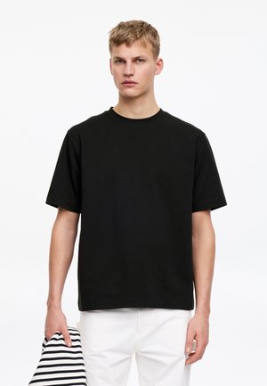 Young man with short blond hair wearing a plain black short-sleeve t-shirt and white pants, holding folded striped fabric in left hand.