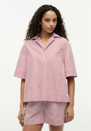Jonge vrouw die een rood-wit verticaal gestreept kortärmelig shirt en bijpassende korte broek draagt, staand tegen een effen achtergrond.