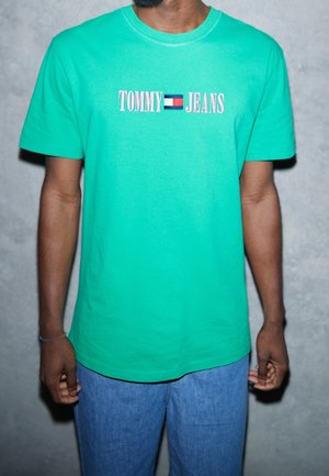 Uomo che indossa una maglietta verde brillante di Tommy Jeans con testo rosa e un piccolo logo rosso, bianco e blu, abbinata a jeans blu.