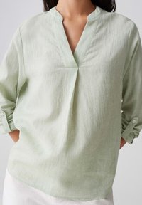 Blusa in lino verde chiaro con scollo a V, maniche lunghe arrotolate al gomito e vestibilità morbida, caratterizzata da una sottile texture e dettagli a pieghe.