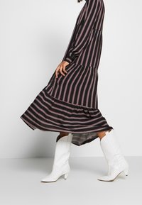 Černé maxi šaty s vertikálními pruhy v růžové a bílé barvě, ve vrstveném designu, doplněné bílými, texturovanými botami do půli lýtek s špičatou špičkou.