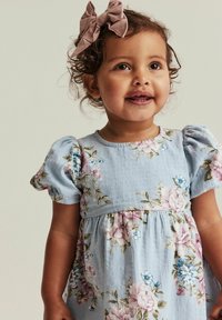 Taivasinsininen puuvillamekko, jossa on vaaleanpunaisia ja sinisiä kukkakuoseja. Puffihihat ja kerätty vyötärö. Ruskeat hiukset, joissa on vaaleanpunainen nauha.