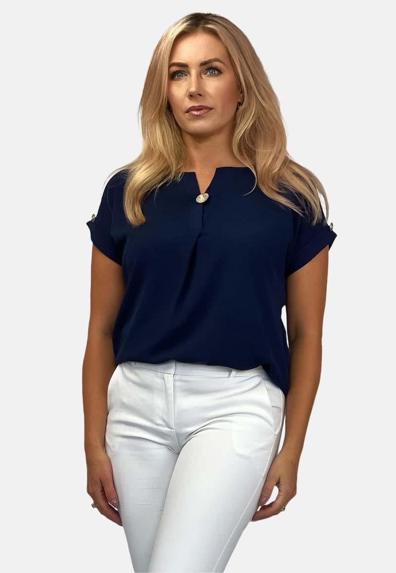 Granatowa bluzka z krótkim rękawem z detalem w postaci guzika przy dekolcie i zawiniętymi mankietami, zestawiona z białymi dopasowanymi spodniami. Gładka struktura.