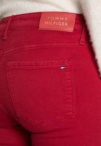 Detalle de unos pantalones vaqueros rojos de Tommy Hilfiger, mostrando la etiqueta de la marca encima del bolsillo trasero y la pequeña bandera del logo en la esquina del bolsillo.