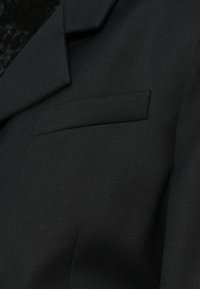 Musta bleiser, millel on tekstureeritud kangas, sisselõigatud kraed ja üks rinna tasku. Lihtne ja struktureeritud disain puhaste joontega.