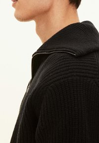 Svart stickad tröja med en strukturerad yta, med en hög krage och en dragkedja längs framsidan. Närbild av halsringningen.