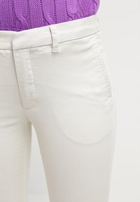 Ljust beige bomullsblandade byxor med en slät yta, som har midjeband och framfickor, ihop med en texturerad lila tröja.