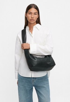 Donna che indossa una camicia bianca con bottoni e jeans blu, mentre tiene una borsa a tracolla in pelle nera con un logo visibile sul davanti.