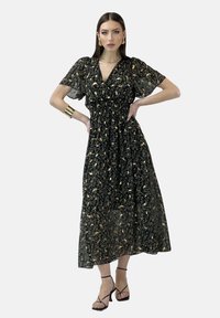 Robe maxi noire à motif floral, avec de courtes manches volantées, taille froncée et texture transparente. Des accents dorés dans le motif floral ajoutent des détails.