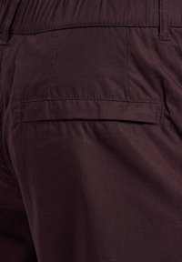 Donkerbordeauxrode broek met een gladde textuur, voorzien van een achterzak zonder zichtbare hardware of versieringen. Subtiele stiknaaddetails.